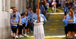 A priest is carrying a cross down the flowered streets of Cagli. Behind him are scouts. The scouts come early to help decorate the streets in honor of the procession.